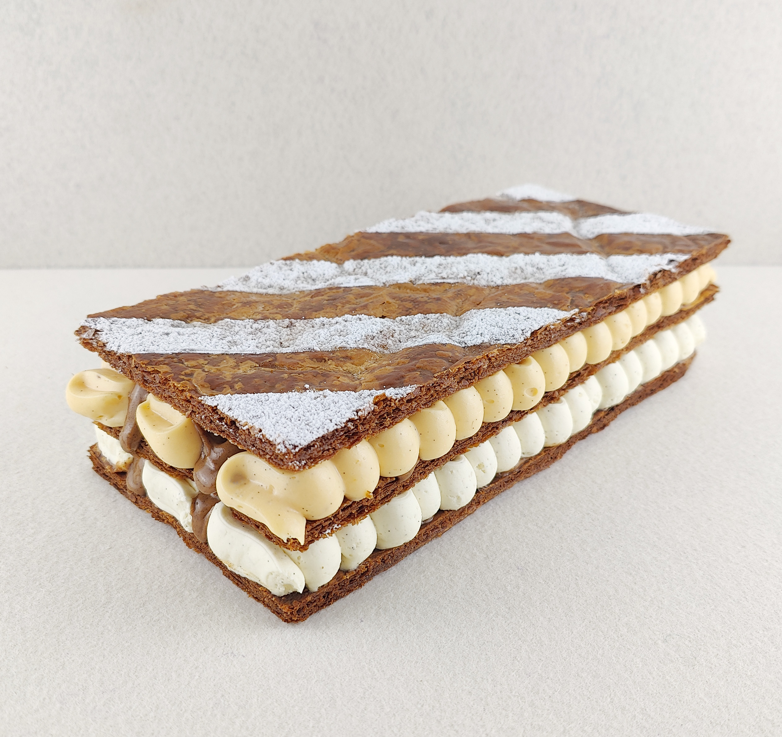 Milhojas rectangular para varias personas fotografiado en ángulo de 45 grados sobre fondo claro, con tres capas de hojaldre, rellenos de ganache montada de chocolate blanco, vainilla y mascarpone, muselina ligera de vainilla y decoración superior de franjas alternas de hojaldre y azúcar glas.