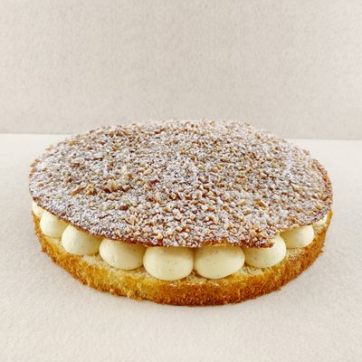 Tarta Tropézienne grande para compartir, fotografiada en ángulo de 45 grados, con brioche dorado relleno de crema diplomática a la vainilla y superficie cubierta de azúcar perlado, almendra picada y azúcar glas, sobre fondo vacío y de color blanco roto.