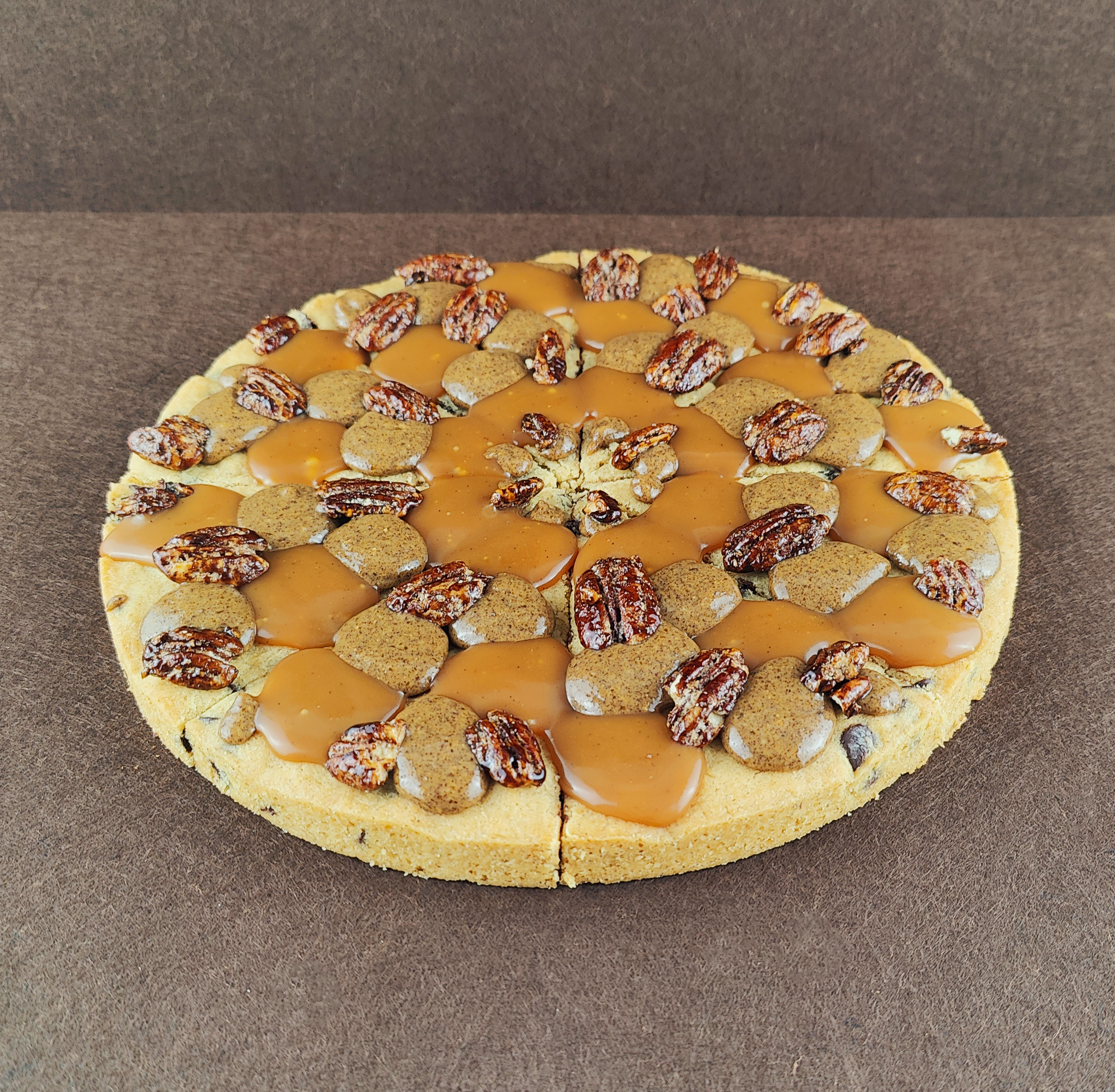 Grand Cookie de pecana vista en ángulo de 45 grados sobre fondo marrón claro, con chips de chocolate negro, tofe de vainilla, praliné de pecana y pecanas garrapiñadas.