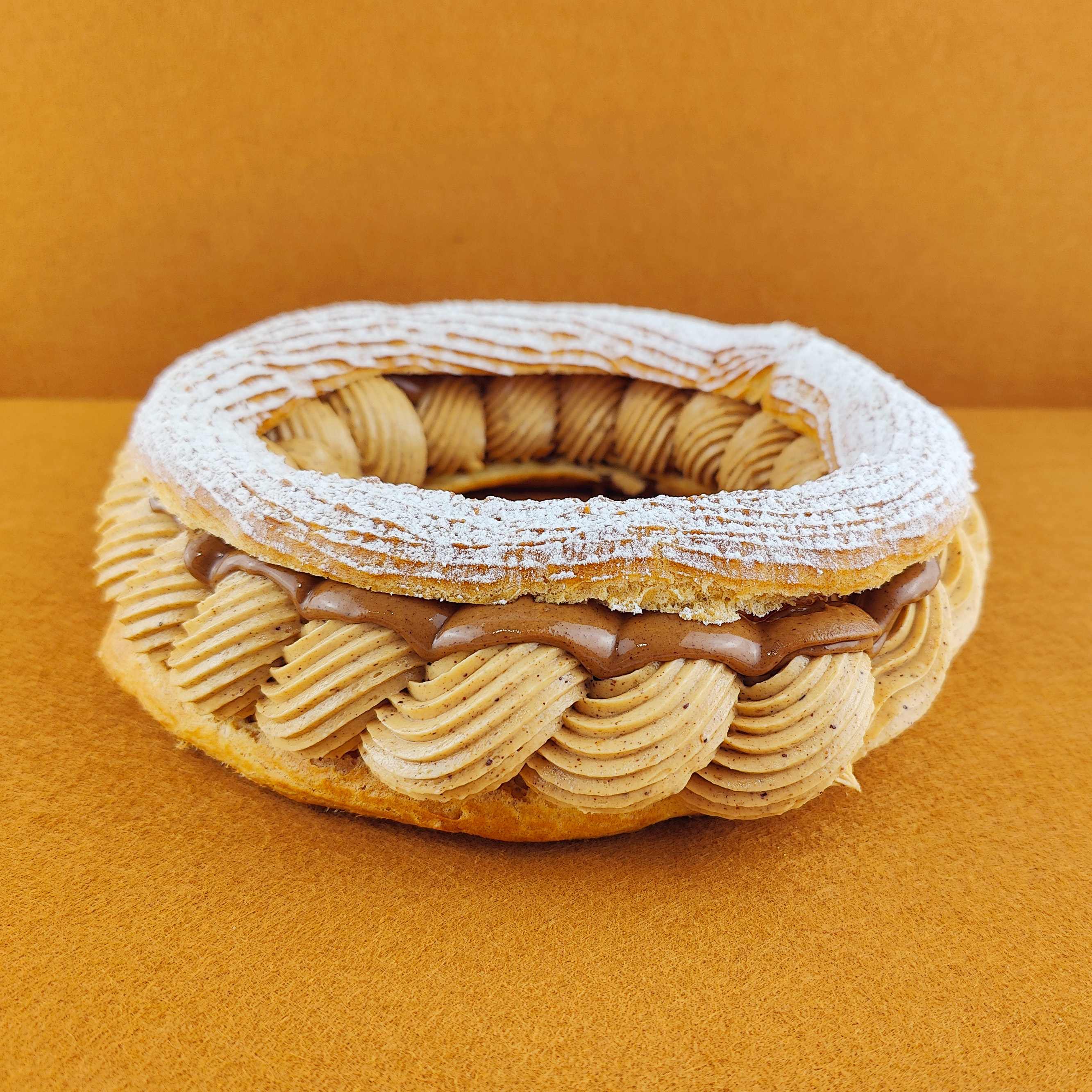 Tarta París-Brest fotografiada en ángulo de 45 grados sobre fondo vacío de color marrón claro, con anillo de masa choux relleno de crema muselina de praliné y praliné por encima, decorada con azúcar glas sobre la parte superior del anillo de masa choux.