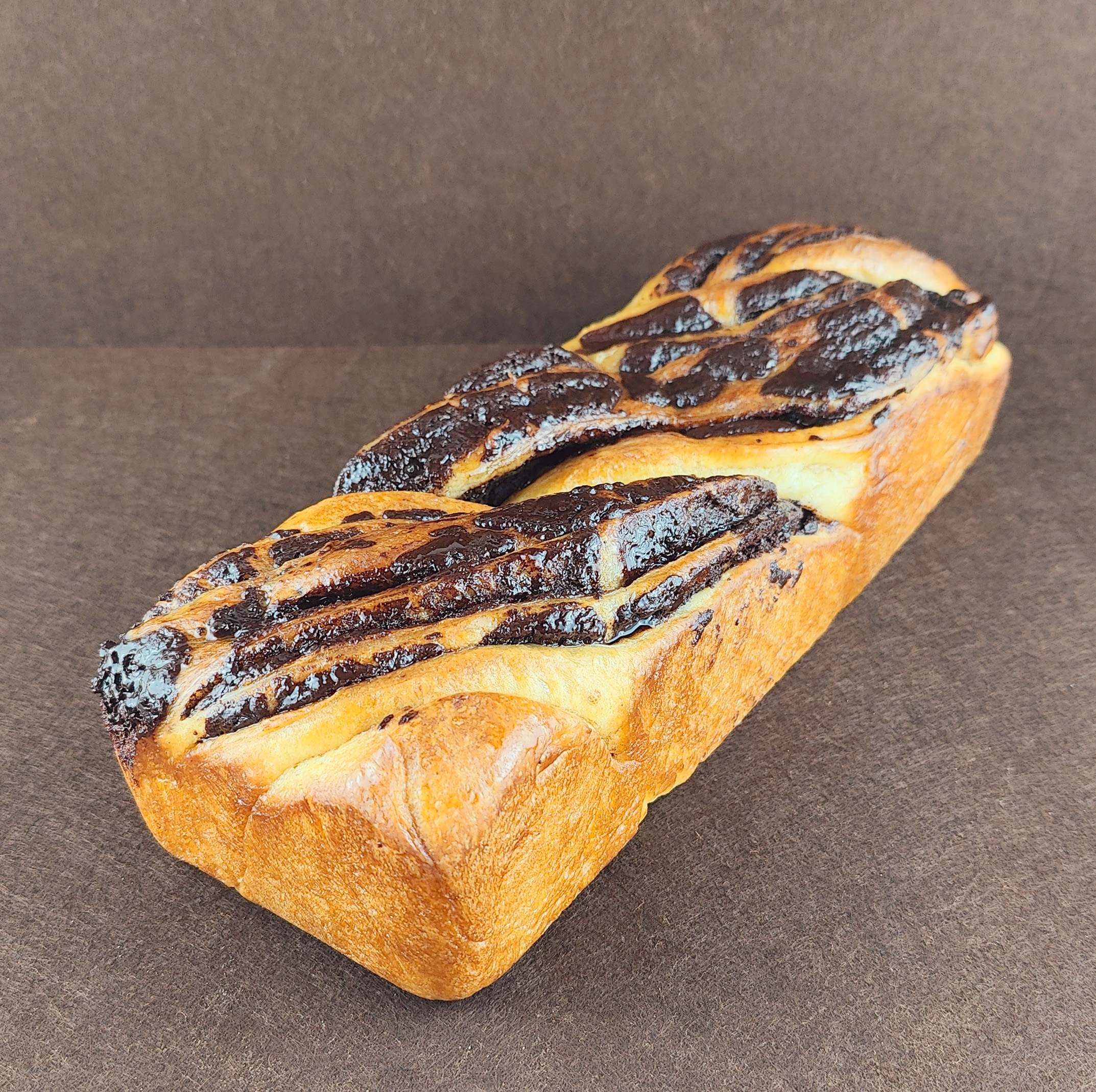 Babka de chocolate fotografiada en ángulo de 45 grados sobre fondo marrón claro, con masa de brioche trenzada, relleno de crema de chocolate y superficie ligeramente glaseada.