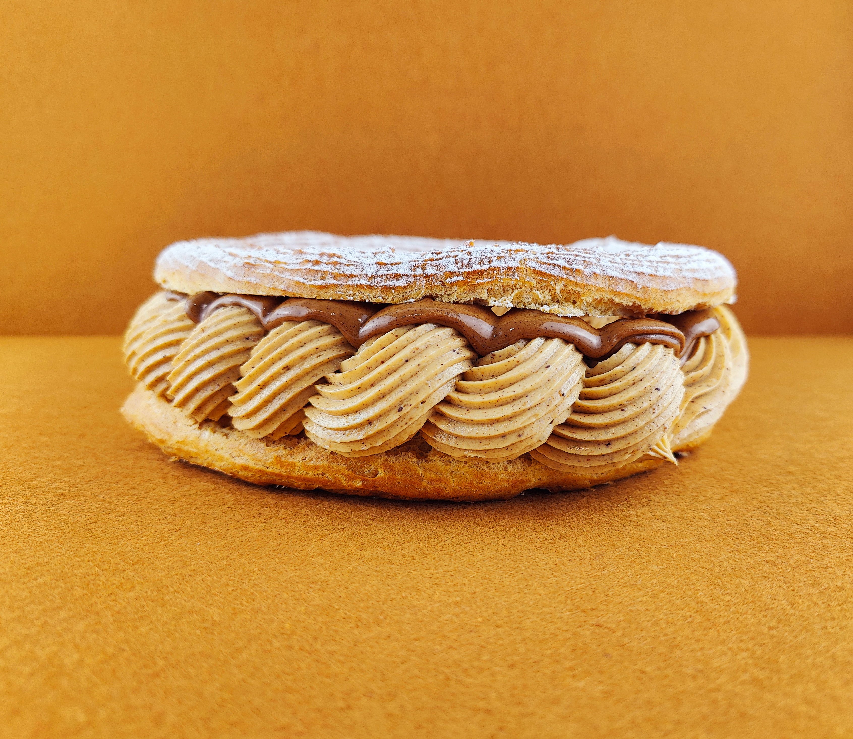Tarta París-Brest vista de frente sobre fondo vacío de color marrón claro, mostrando el anillo de masa choux relleno de crema muselina de praliné con praliné encima, decorada con azúcar glas sobre la parte superior del anillo de masa choux.