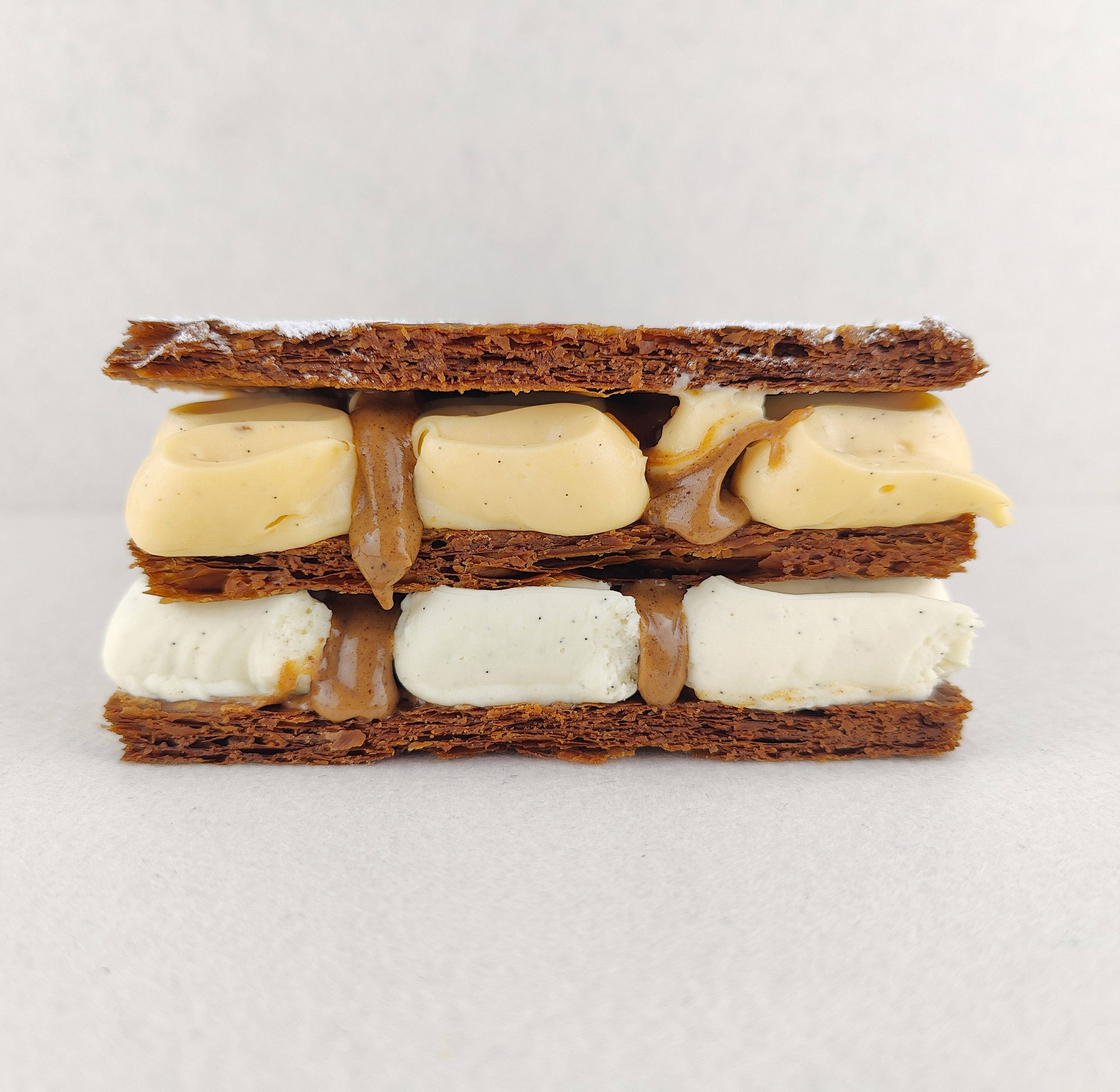 Milhojas rectangular visto de frente, mostrando las tres capas de hojaldre, la ganache montada de chocolate blanco, vainilla y mascarpone en la parte inferior, la muselina ligera de vainilla en la parte superior y dos hilos de praliné en cada relleno.