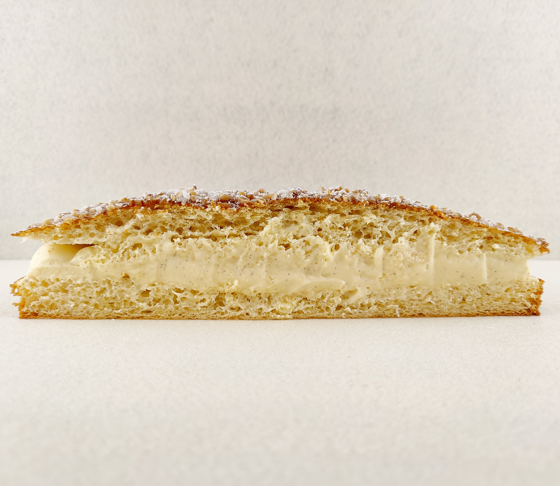 Tarta Tropézienne grande cortada a la mitad y fotografiada de frente, dejando ver el relleno de crema diplomática a la vainilla entre las capas de brioche, con la superficie espolvoreada de azúcar perlado, almendra picada y azúcar glas, sobre fondo vacío y de color blanco roto.