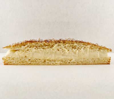 Tarta Tropézienne grande cortada a la mitad y fotografiada de frente, dejando ver el relleno de crema diplomática a la vainilla entre las capas de brioche, con la superficie espolvoreada de azúcar perlado, almendra picada y azúcar glas, sobre fondo vacío y de color blanco roto.