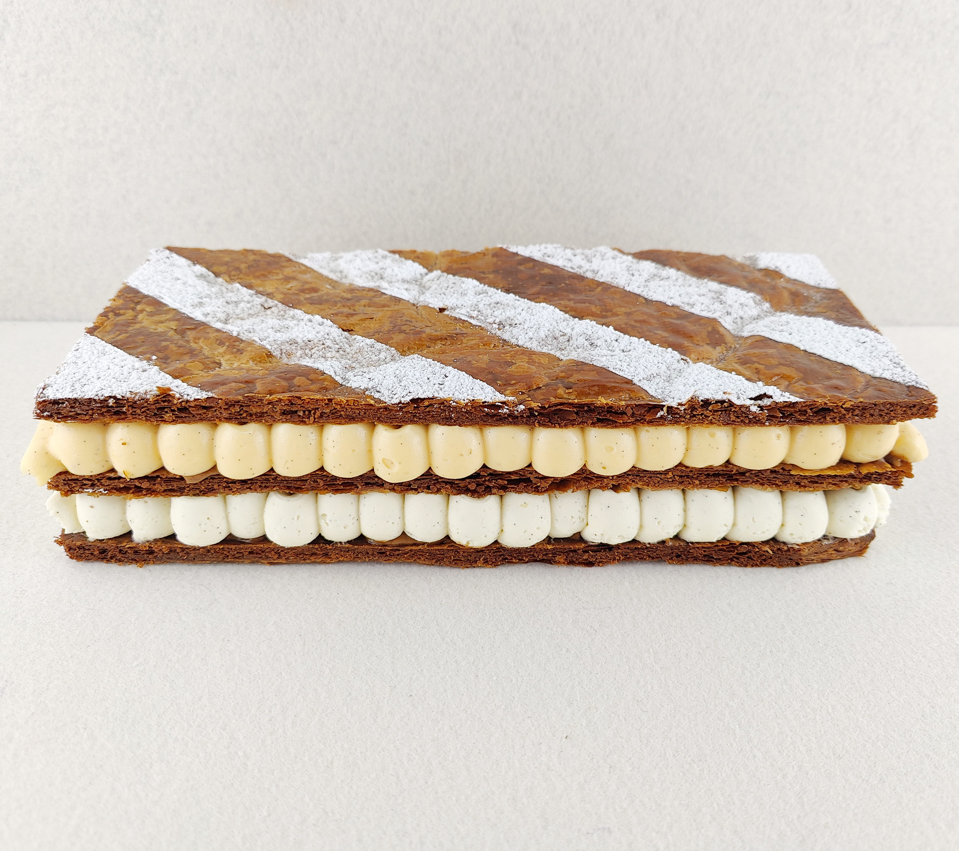 Milhojas rectangular fotografiado en perspectiva lateral de 45 grados, con tres capas de hojaldre crujiente, rellenos de vainilla, mascarpone y praliné, y superficie decorada con franjas diagonales de azúcar glas.