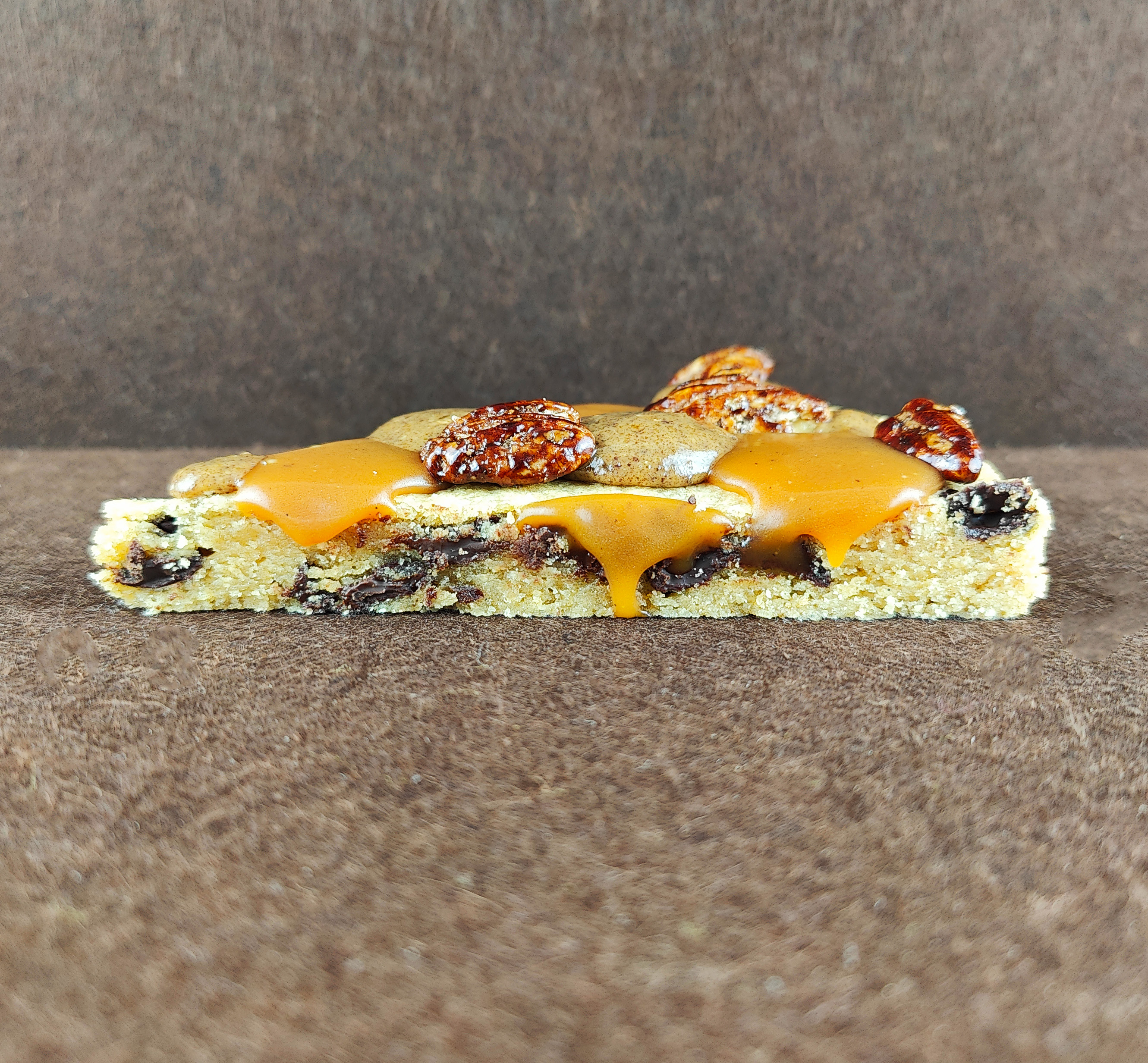 Porción de Grand Cookie de pecana sobre fondo marrón claro, con el interior tierno de la cookie, chips de chocolate negro, tofe de vainilla, praliné de pecana y pecanas garrapiñadas.
