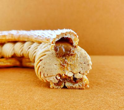Tarta Paris-Brest cortada a la mitad y fotografiada de frente sobre fondo vacío de color marrón claro, mostrando el interior del anillo de masa choux con avellanas garrapiñadas y praliné en la base, crema muselina de praliné, y más praliné cayendo suntuosamente desde la parte superior, con azúcar glas decorando la superficie del anillo superior.
