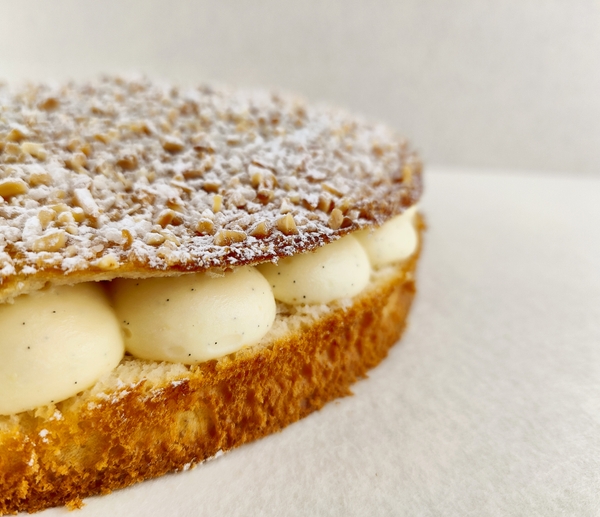 Primer plano de la Tarta Tropézienne, donde se aprecian las texturas de la crema diplomática a la vainilla en el interior, la miga esponjosa del brioche y la cubierta con azúcar perlado, almendra picada y azúcar glas, sobre fondo vacío y de color blanco roto.