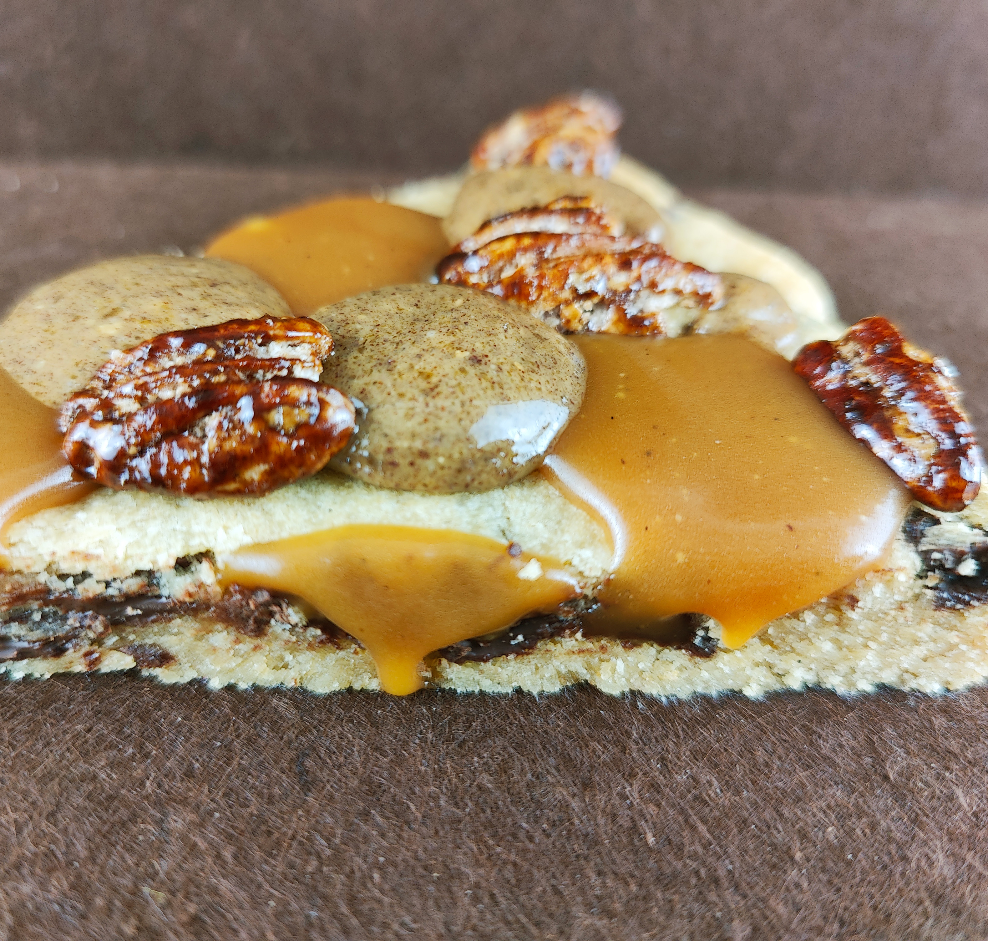 Detalle cercano de la Grand Cookie de pecana sobre fondo marrón claro, resaltando el tofe de vainilla, el praliné de pecana y las pecanas garrapiñadas de la superficie.