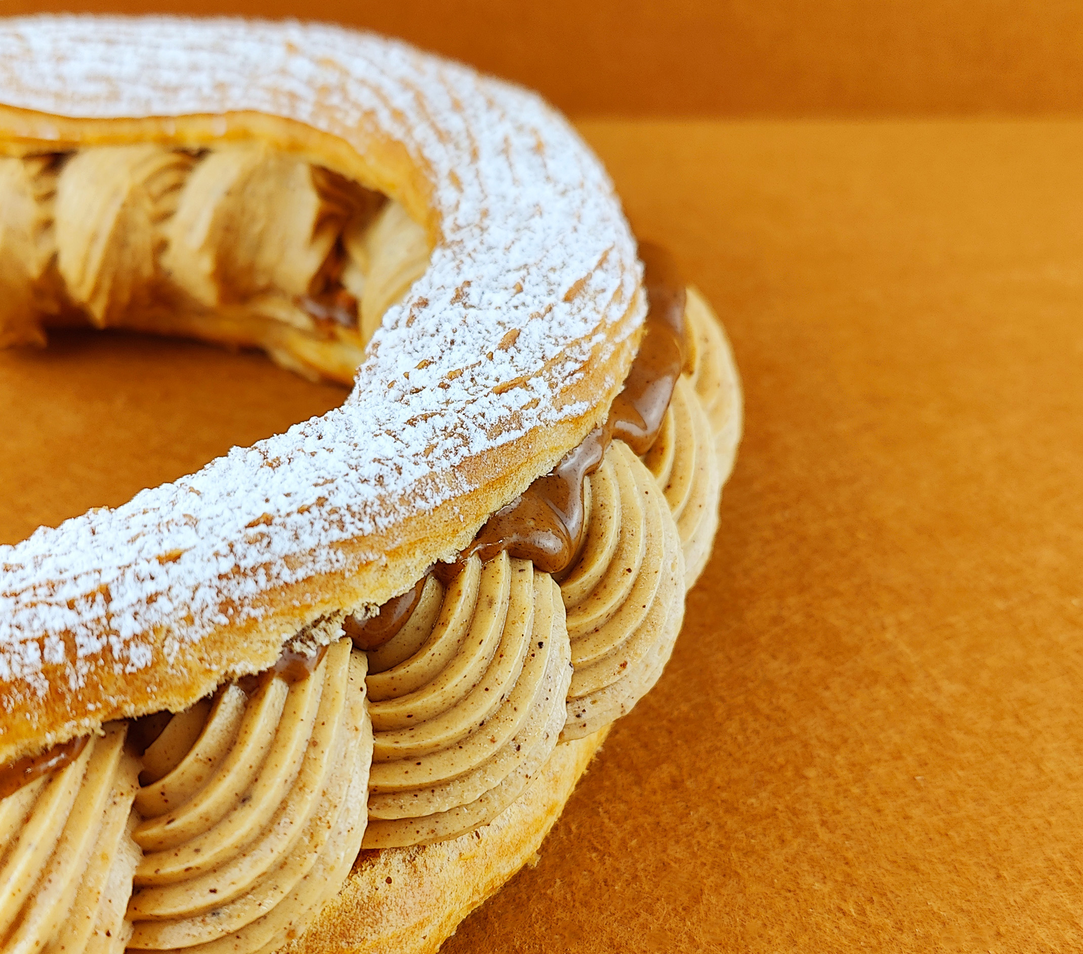 Primer plano de Tarta París-Brest sobre fondo vacío de color marrón claro, resaltando la textura de la crema muselina de praliné, el brillo del praliné y la cobertura de azúcar glas sobre el anillo de masa choux.