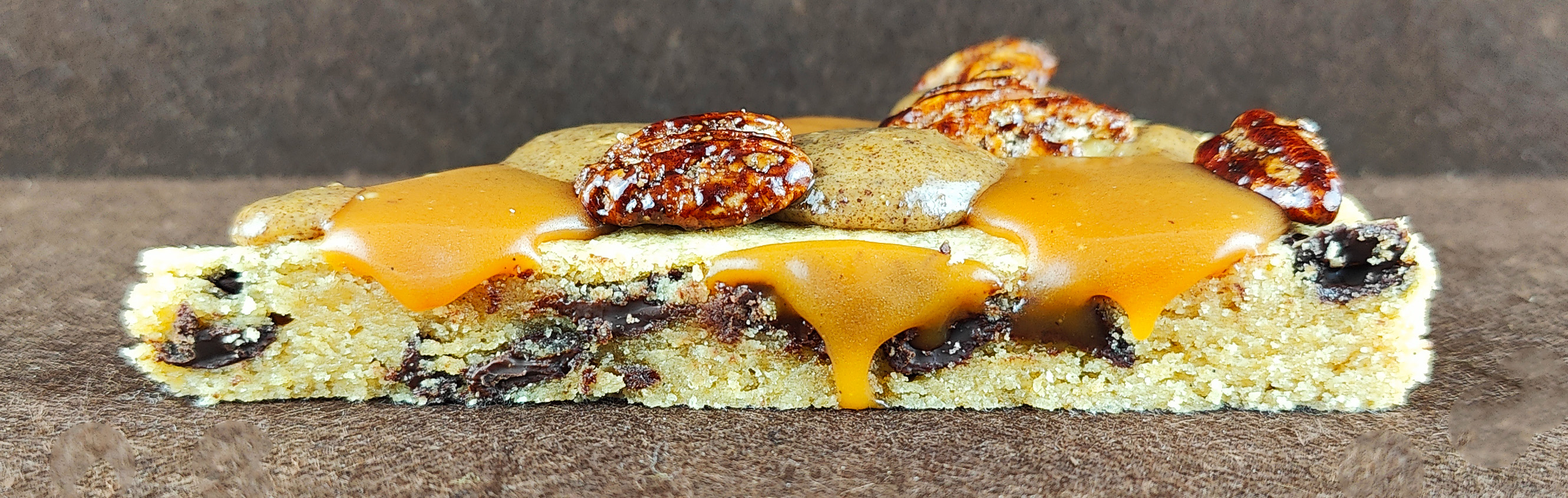 Porción de Grand Cookie de pecana sobre fondo marrón claro, con el interior tierno de la cookie, chips de chocolate negro, tofe de vainilla, praliné de pecana y pecanas garrapiñadas.