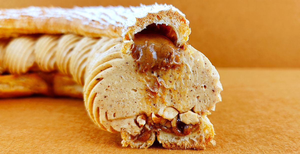 Tarta Paris-Brest individual cortada a la mitad y fotografiada de frente sobre fondo vacío de color marrón claro, mostrando el interior del anillo de masa choux con avellanas garrapiñadas y praliné en la base, crema muselina de praliné, y más praliné cayendo suntuosamente desde la parte superior, con azúcar glas decorando la superficie del anillo superior.