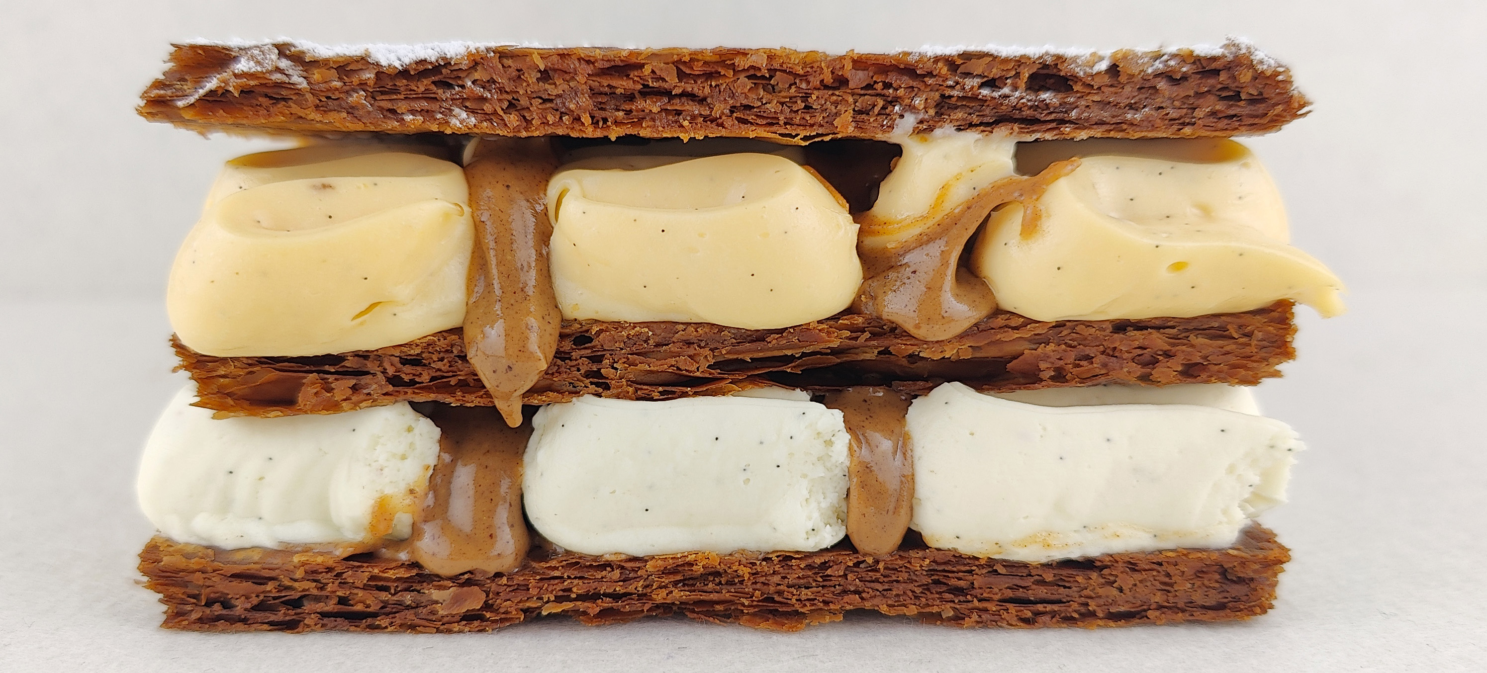 Milhojas rectangular fotografiado en perspectiva lateral de 45 grados, con tres capas de hojaldre crujiente, rellenos de vainilla, mascarpone y praliné, y superficie decorada con franjas diagonales de azúcar glas.