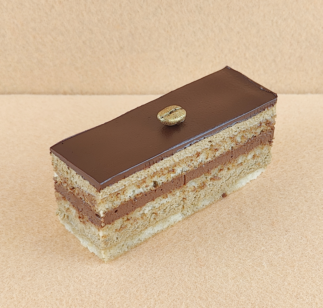 Porción individual rectangular de Tarta Ópera, fotografiada en ángulo de 45 grados con ligera inclinación, mostrando la cobertura de chocolate lisa y un grano de café decorativo en la parte superior.