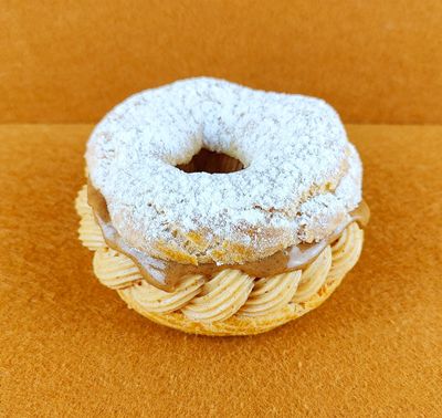 Tarta Paris-Brest individual fotografiada en ángulo de 45 grados sobre fondo vacío de color marrón claro, con anillo de masa choux relleno de crema muselina de praliné y praliné por encima, decorada con azúcar glas sobre la parte superior del anillo de masa choux.