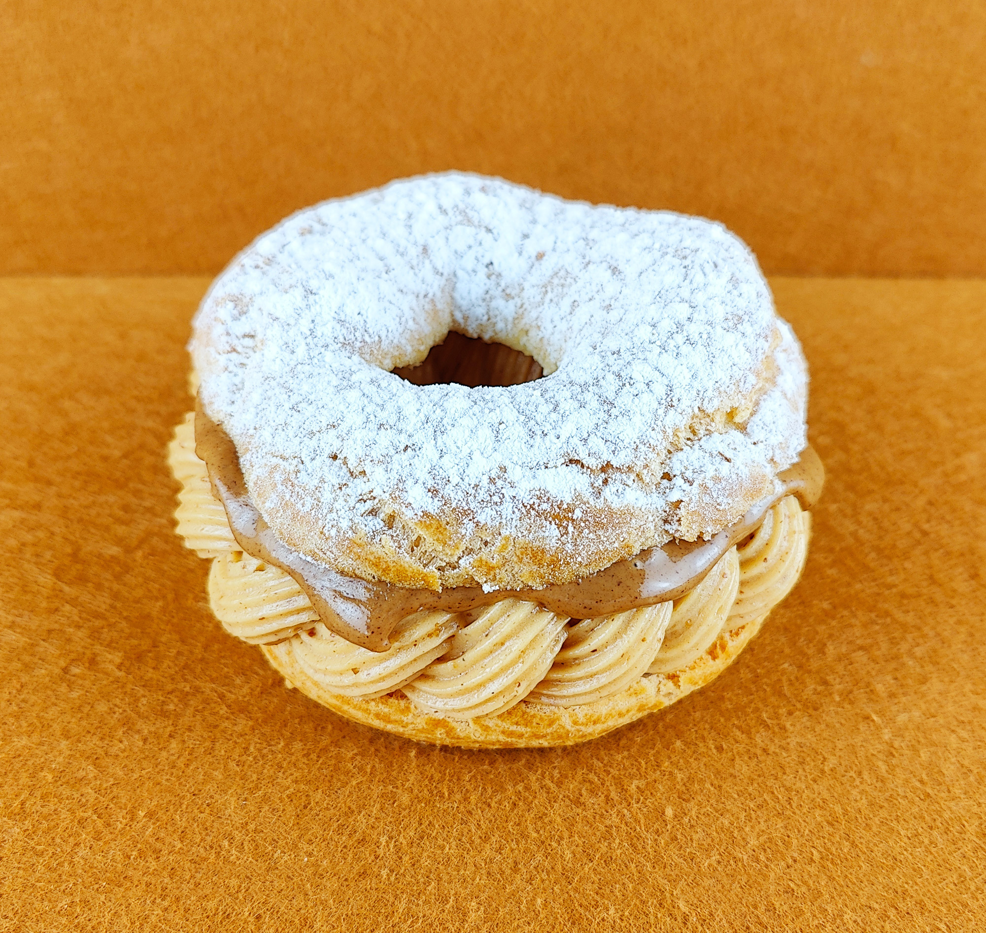 Tarta París-Brest individual fotografiada en ángulo de 45 grados sobre fondo vacío de color marrón claro, con anillo de masa choux relleno de crema muselina de praliné y praliné por encima, decorada con azúcar glas sobre la parte superior del anillo de masa choux.