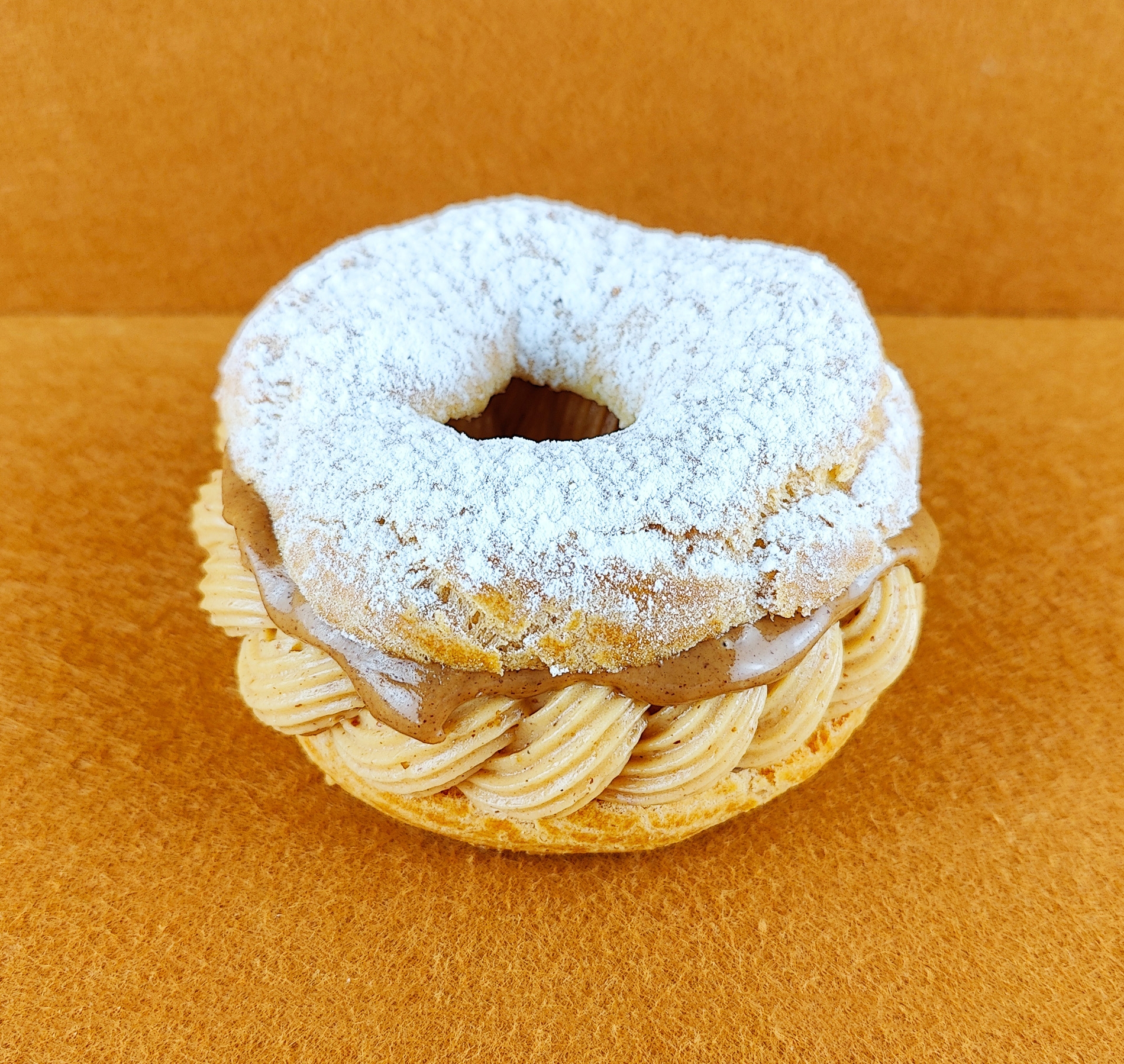 Tarta Paris-Brest individual fotografiada en ángulo de 45 grados sobre fondo vacío de color marrón claro, con anillo de masa choux relleno de crema muselina de praliné y praliné por encima, decorada con azúcar glas sobre la parte superior del anillo de masa choux.
