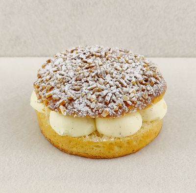 Primer plano de la Tarta Tropézienne, donde se aprecian las texturas de la crema diplomática a la vainilla en el interior, la miga esponjosa del brioche y la cubierta con azúcar perlado, almendra picada y azúcar glas, sobre fondo vacío y de color blanco roto.