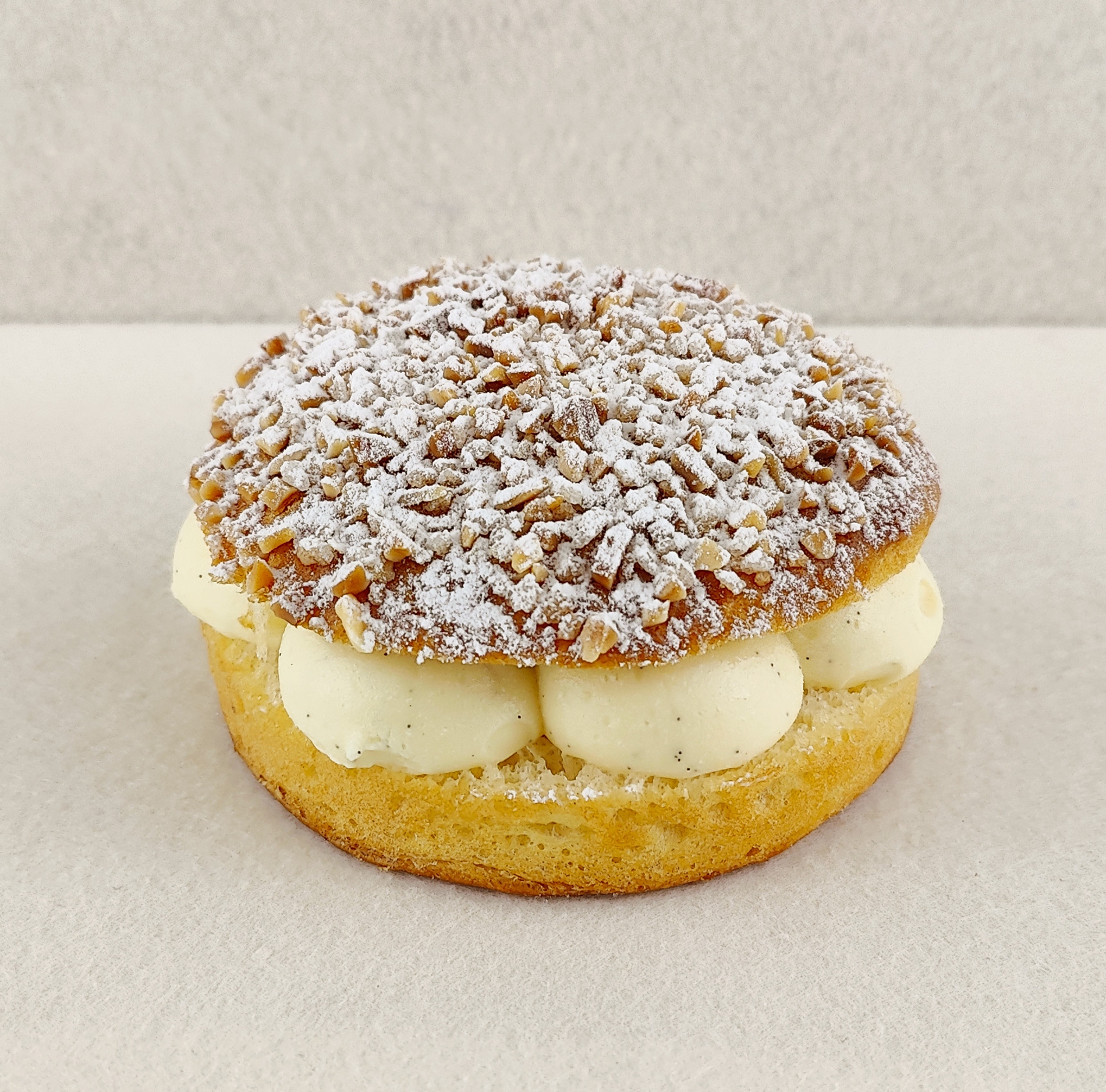 Primer plano de la Tarta Tropézienne, donde se aprecian las texturas de la crema diplomática a la vainilla en el interior, la miga esponjosa del brioche y la cubierta con azúcar perlado, almendra picada y azúcar glas, sobre fondo vacío y de color blanco roto.
