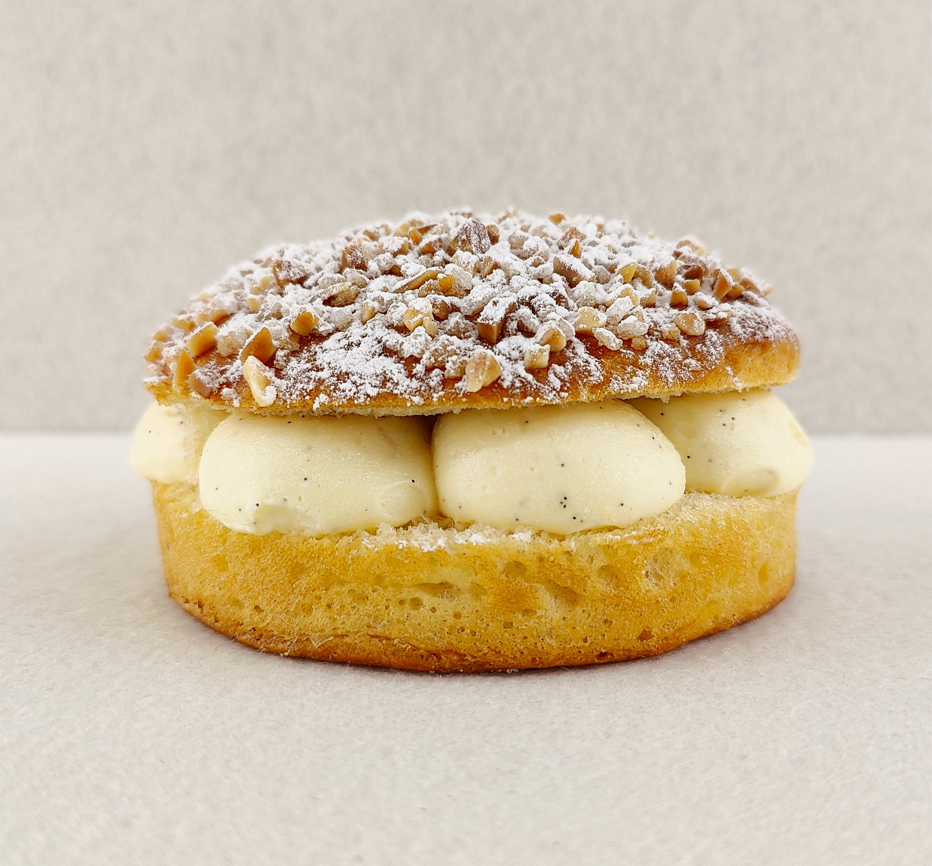 Tarta Tropézienne pequeña individual vista de frente, mostrando el brioche dorado relleno de crema diplomática a la vainilla, con la parte superior cubierta de azúcar perlado, almendra picada y azúcar glas, sobre fondo vacío y de color blanco roto.