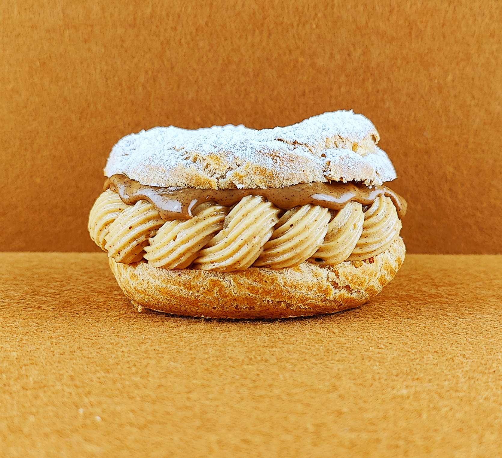 Tarta París-Brest individual vista de frente sobre fondo vacío de color marrón claro, mostrando el anillo de masa choux relleno de crema muselina de praliné con praliné encima, decorada con azúcar glas sobre la parte superior del anillo de masa choux.