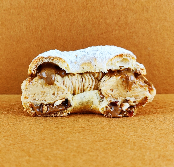 Tarta Paris-Brest individual cortada a la mitad y fotografiada de frente sobre fondo vacío de color marrón claro, mostrando el interior del anillo de masa choux con avellanas garrapiñadas y praliné en la base, crema muselina de praliné, y más praliné cayendo suntuosamente desde la parte superior, con azúcar glas decorando la superficie del anillo superior.