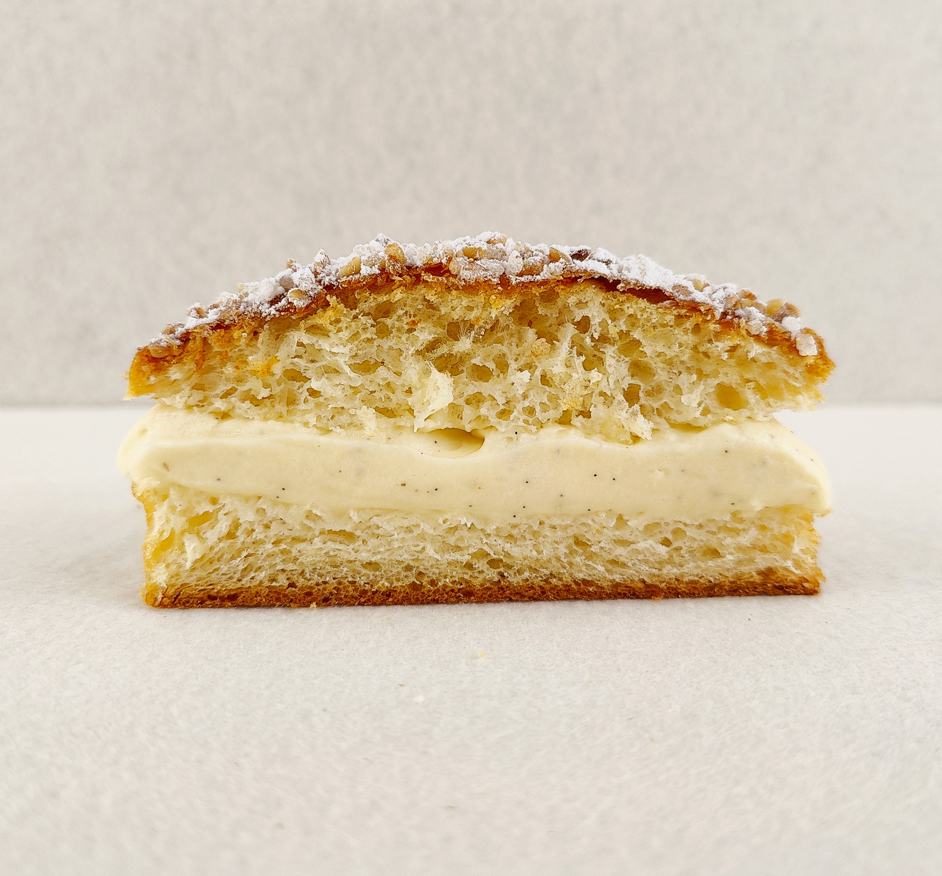 Primer plano de Tarta Tropézienne pequeña individual, donde se aprecian las texturas de la crema diplomática a la vainilla en el interior, la miga esponjosa del brioche y la cubierta con azúcar perlado, almendra picada y azúcar glas, sobre fondo vacío y de color blanco roto.