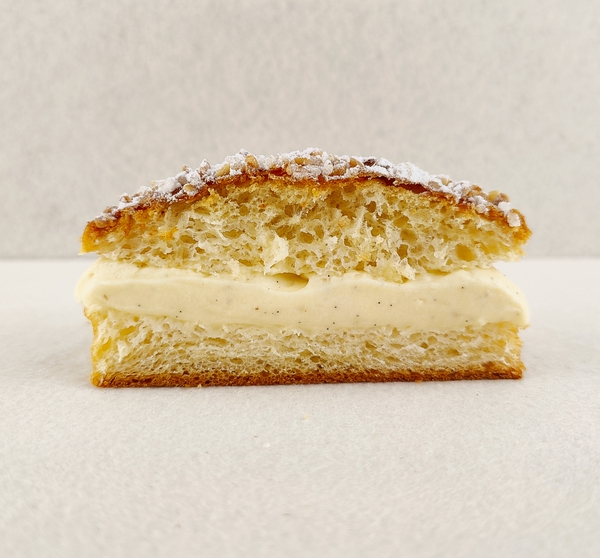 Primer plano de Tarta Tropézienne pequeña individual, donde se aprecian las texturas de la crema diplomática a la vainilla en el interior, la miga esponjosa del brioche y la cubierta con azúcar perlado, almendra picada y azúcar glas, sobre fondo vacío y de color blanco roto.